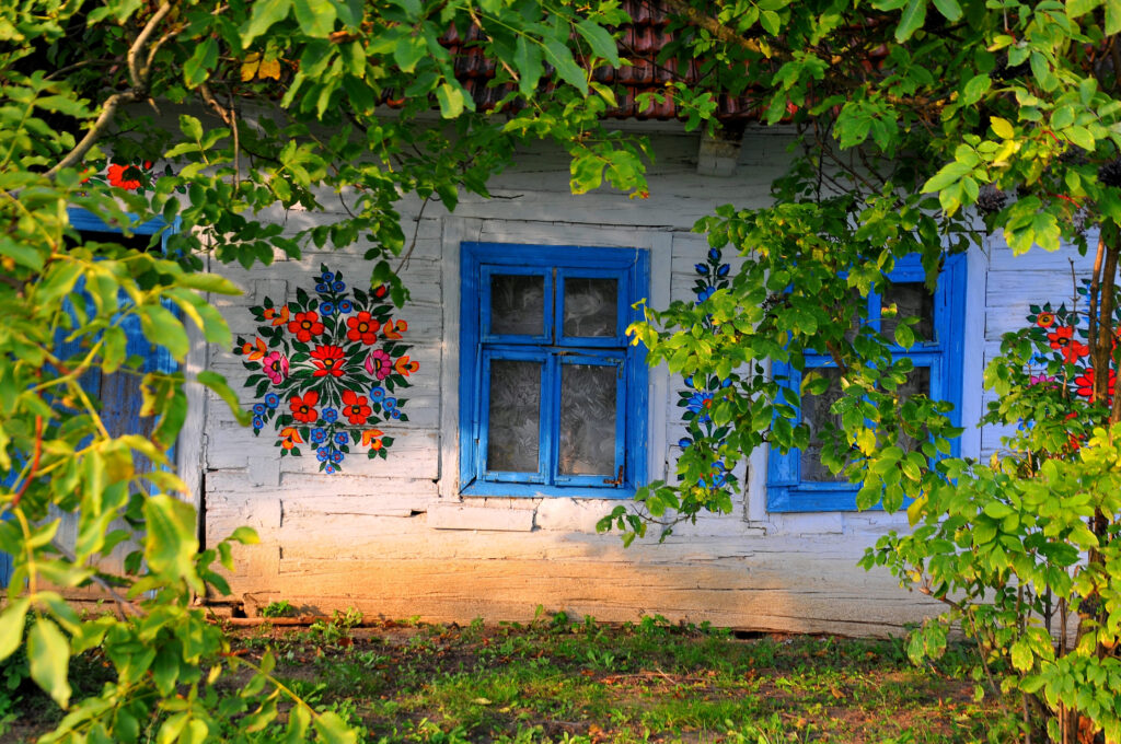 Ściana wiejskiego domu w Zalipiu z niebieskim oknem otoczona kwiatami i malunkami